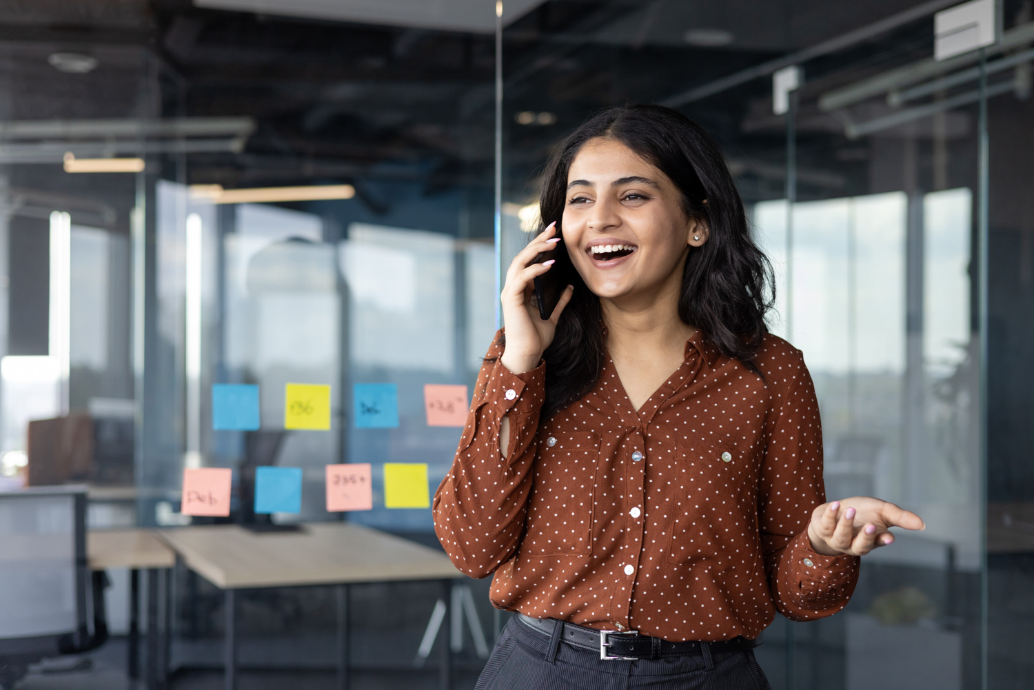 successful and happy office worker talking on the phone while standing near the window inside the office building. satisfied businesswoman at workplace communicating with colleagues and clients